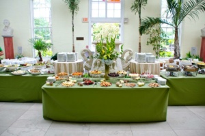 An elegant buffet setup with tables covered in green tablecloths, displaying a variety of dishes and desserts. The backdrop includes tall windows, plants, and decorative sculptures. Plates are stacked on tables, ready for serving.