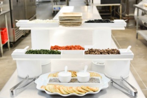 A kitchen with a display of ingredients, including small trays of chopped vegetables, herbs, and toppings, placed on a multi-tiered stand. In the foreground, a tray of thinly sliced food with small bowls for dipping and utensils.