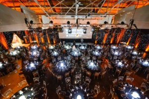 A large, dimly lit event hall with round tables set for a formal gathering. Warm orange and white lighting illuminates the space. A stage at the front displays a presentation screen, and decorative lights hang overhead.