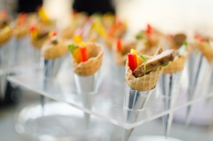 A display of small waffle cones filled with colorful ingredients like red and yellow bell peppers, garnished with herbs, and held upright in silver stands. The image has a blurred background, focusing on the vibrant appetizers.