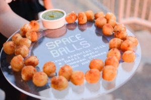 A circular tray of small, golden-brown fried balls arranged around a sauce dish with green pesto. The tray reads "SPACE BALLS - citrus arancini with a basil pesto and parmesan cheese and pine nuts.