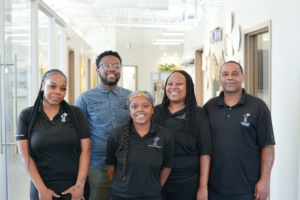 A group of five people standing indoors, smiling at the camera. Four wear matching black polo shirts, and one wears a blue button-up shirt. The room has bright lighting and visible office decor in the background.