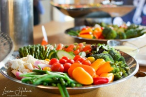 A colorful vegetable platter featuring broccoli, cherry tomatoes, bell peppers, radishes, cucumbers, and asparagus on a wooden table, with a bowl of dressing in the background. The setting suggests a buffet or event.