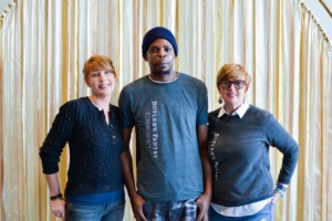 Three people stand in front of a gold curtain. The person in the middle wears a beanie and gray shirt, while the two women on either side wear glasses and casual shirts. The shirts read "Butler's Pantry." They are smiling casually.