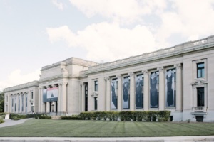 Exterior view of a large, classical-style museum building with tall columns and large banners hanging between the windows. The sky is partly cloudy, and a well-maintained lawn is in the foreground.