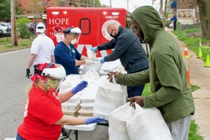 Volunteers distribute food to people at a community event. They are wearing masks and gloves, with a red emergency vehicle in the background. Several individuals are accepting bags from a table set up on the street.