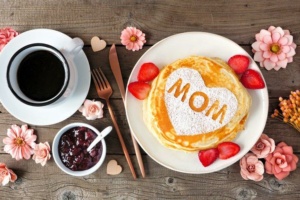 A table setting with a cup of coffee, jam, and pancakes topped with a heart-shaped powdered sugar design with "MOM" written on it. Strawberries and pink flowers decorate the setting.