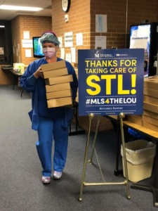 Healthcare worker in blue scrubs and mask holding several stacked boxes. Next to them is a sign reading, "Thanks for taking care of STL! #MLS4THELOU." The setting appears to be inside a hospital or clinic.