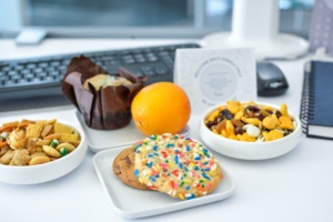 An office desk with snacks: a muffin in a brown wrapper, an orange, a plate of a sprinkle-covered cookie, a bowl of mixed nuts and raisins, and a bowl with savory snack mix. A computer keyboard, mouse, and notebook are in the background.