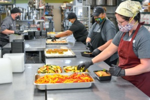 A team of four people in a commercial kitchen preparing and packaging meals. They are wearing masks and gloves, and the meals include colorful vegetables and protein. The kitchen is equipped with stainless steel tables and industrial cookware.