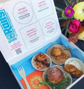 A tasting box containing a chicken "lollipop" with salad, a lobster corn dog with mango chutney, a Pretzel & Draytwerst Kolache with beer mustard and arugula, and a pumpkin funnel cake waffle with spiced cream cheese is displayed. Flowers are nearby.