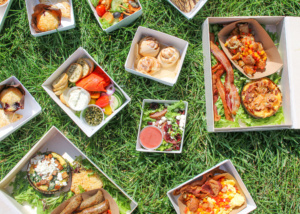 An assortment of picnic food boxes on grass, including quiche, bacon, salad, pastries, and vegetables. The colorful spread is arranged randomly, showcasing a variety of textures and flavors in an outdoor setting.