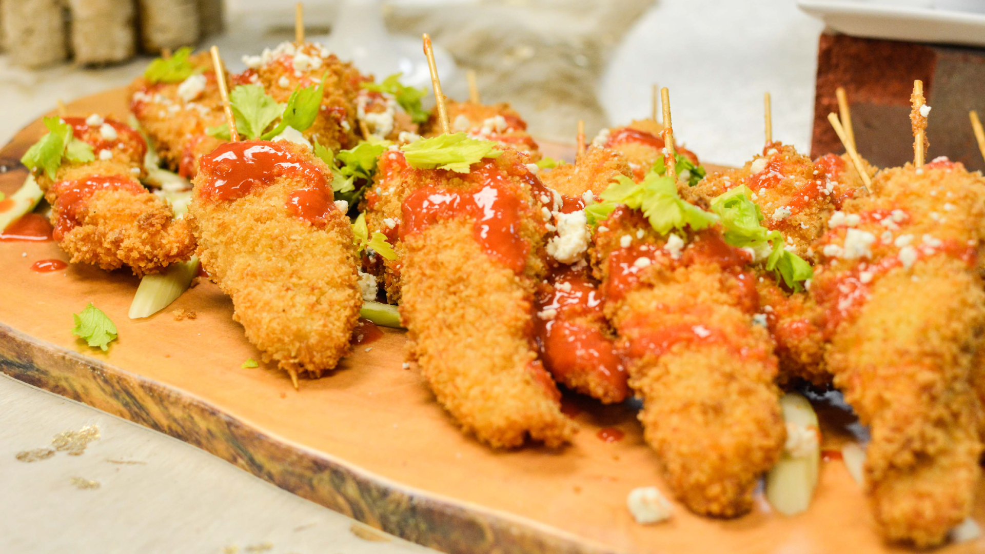 A wooden plate displays golden-brown breaded chicken skewers drizzled with red sauce and garnished with fresh herbs and crumbled cheese.