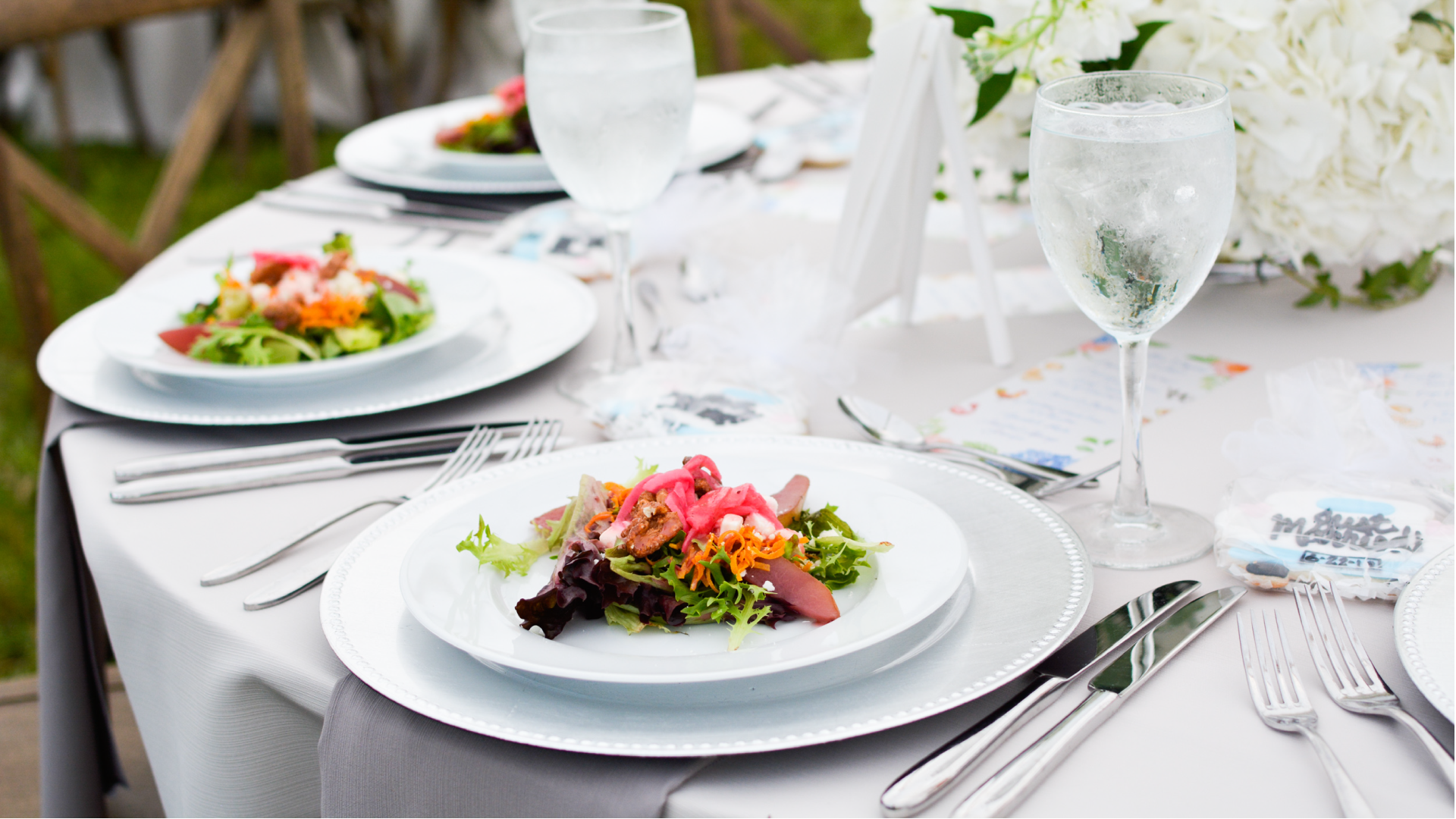 A beautifully set table with a fresh salad on a white plate, accompanied by a glass of water. The table is elegantly arranged with cutlery, a menu, and floral decorations in the background.