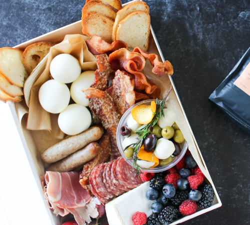 A charcuterie box with boiled eggs, toast, sausages, bacon, prosciutto slices, salami, olives, mozzarella, orange slices, blueberries, raspberries, blackberries, brie cheese, and fresh herbs on a dark surface.