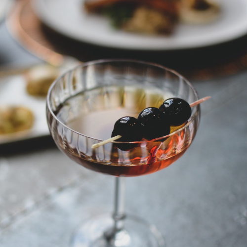 A cocktail in a fluted glass garnished with three dark cherries on a skewer. The background is softly blurred, showing other dishes on a table.