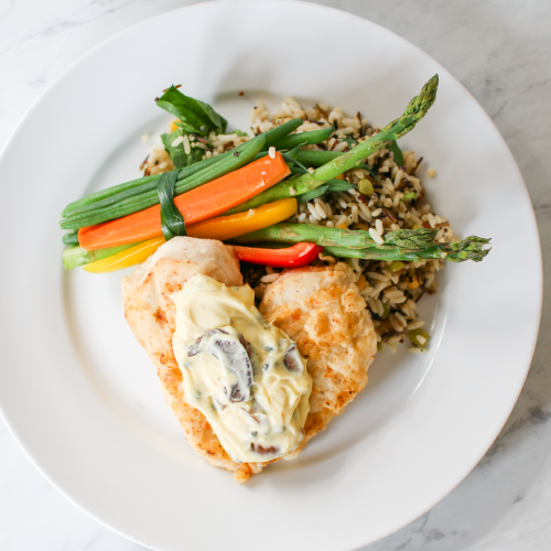 A plate of chicken topped with creamy sauce, served with a bundle of asparagus, carrots, and green beans, tied together with a leek strip, alongside a portion of wild rice on a white plate.
