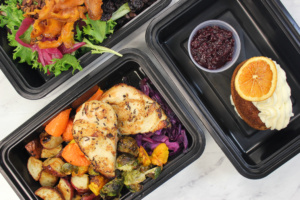 Three black containers with meals: one has grilled chicken, roasted vegetables, and cabbage; another holds a mixed salad with bacon; the third features a cupcake with cream cheese frosting, topped with orange slices, next to a small cup of berry sauce.