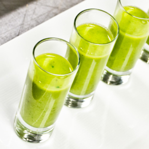 Four small glass tumblers filled with vibrant green smoothie are neatly arranged on a white rectangular tray. The creamy texture and fresh color suggest a blend of fruits or vegetables, potentially served as a refreshing appetizer or health drink.