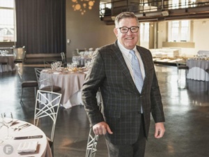 A smiling man in a plaid blazer and glasses stands in an elegant, empty event space. Round tables are set with folded napkins and glassware, ready for a gathering. Soft lighting and large windows create a warm atmosphere.