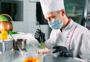 A chef in a white uniform and mask carefully garnishes a dish in a commercial kitchen. He uses tweezers to place herbs on a plated dish, surrounded by various cooking ingredients and kitchen tools.