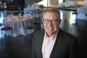A smiling man in glasses and a plaid jacket stands in a room with elegantly set tables. The background shows round tables with white tablecloths and arranged chairs, suggesting a formal event or gathering.