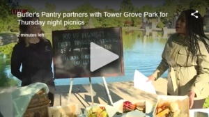 Two people stand by a chalkboard advertising pop-up picnics at Tower Grove Park. A picnic basket and plates of food are on a table in front of them. The scene is set near a lake with trees in the background.