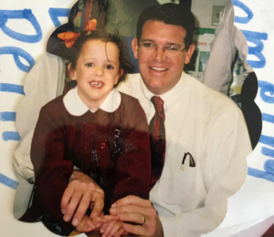 A smiling man in glasses and a tie sits next to a young girl with a large bow in her hair. They are both dressed formally and are sitting in a brightly lit room.