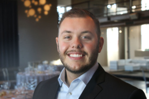 A smiling man in a suit stands in a well-lit room with elegant decor, including round hanging lights and tables set for an event in the background.