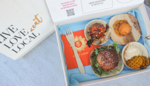 A wooden box open to display a variety of foods: a slider on a bun, a round fried item on a skewer, another round item with sauce, and waffle chips with a dollop of cream. A vibrant red St. Louis napkin is included. The box lid reads "LIVE LOVE LOCAL.