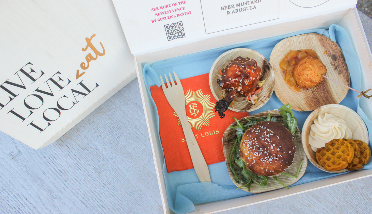 A wooden box open to display a variety of foods: a slider on a bun, a round fried item on a skewer, another round item with sauce, and waffle chips with a dollop of cream. A vibrant red St. Louis napkin is included. The box lid reads "LIVE LOVE LOCAL.
