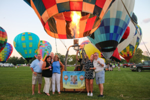 A group of people stands in front of a colorful hot air balloon with flames rising from its burner. More hot air balloons in various colors are visible in the background. The group appears to be celebrating, smiling, and posing for a photo.