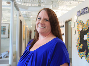 A person with long brown hair and a blue top is smiling while standing in a hallway with art and decorative plates on the walls. The ceiling has pendant lights and the atmosphere is bright.
