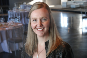 A woman with long blonde hair is smiling, wearing a black leather jacket. She stands in a softly lit room with round tables set with plates and silverware in the background.