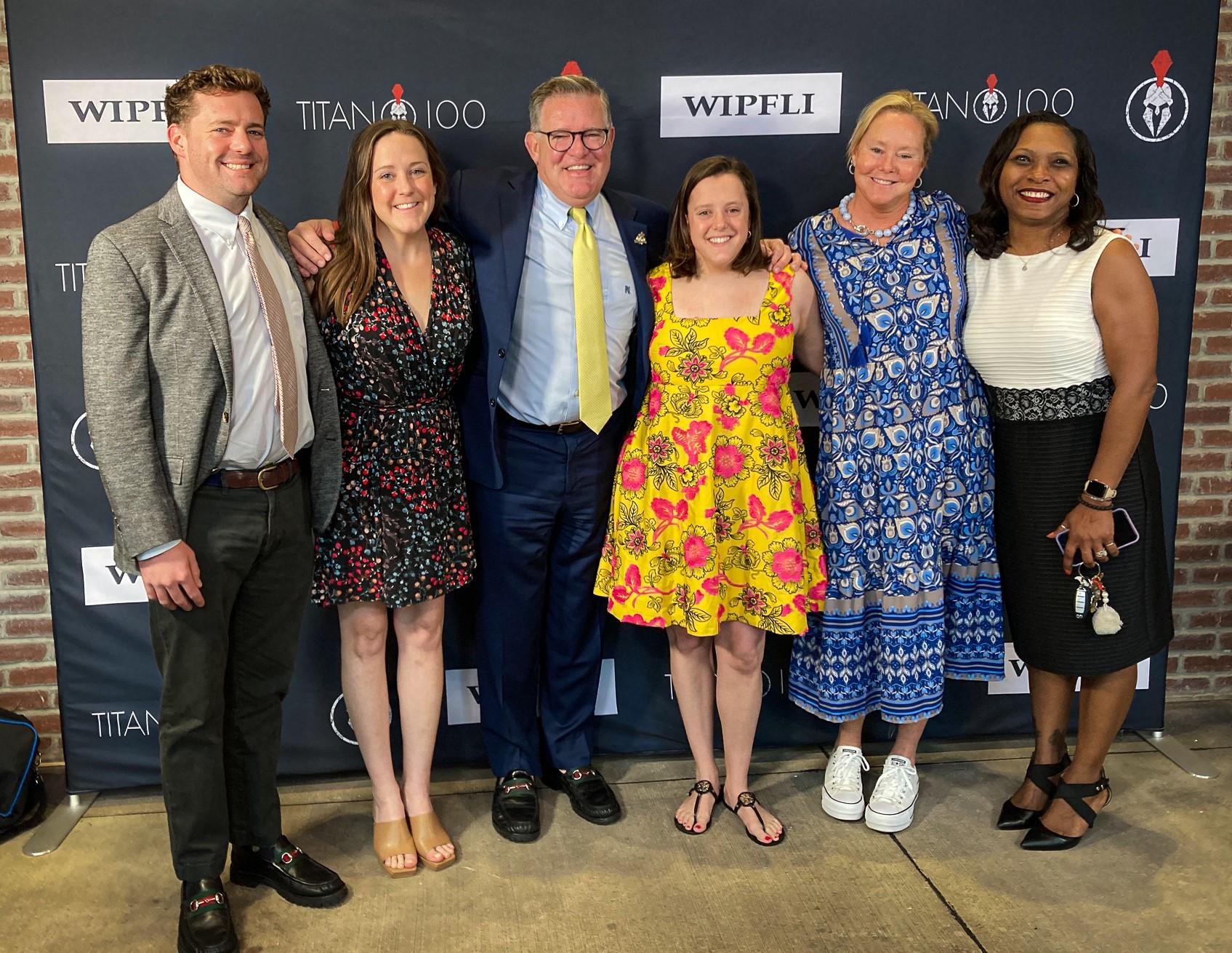 A group of six people standing in front of a backdrop with logos. They are smiling and dressed in formal and semi-formal attire, with men in suits and women in dresses. The setting appears to be at an event or gathering.