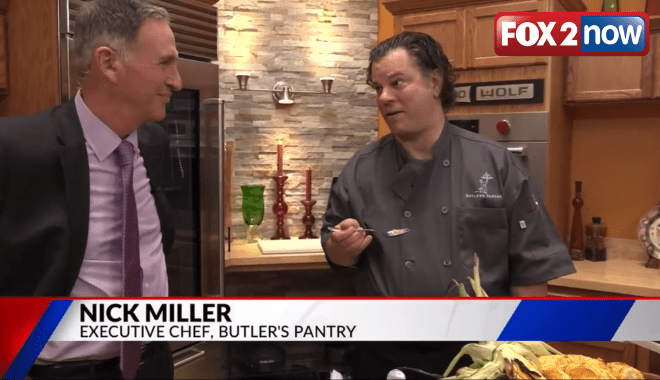 Two men in a kitchen are having a conversation. One, wearing a black chef's coat, holds a spoon and stands next to a table with food. The other, in a suit, listens attentively. A "FOX 2 now" logo is in the top right corner.