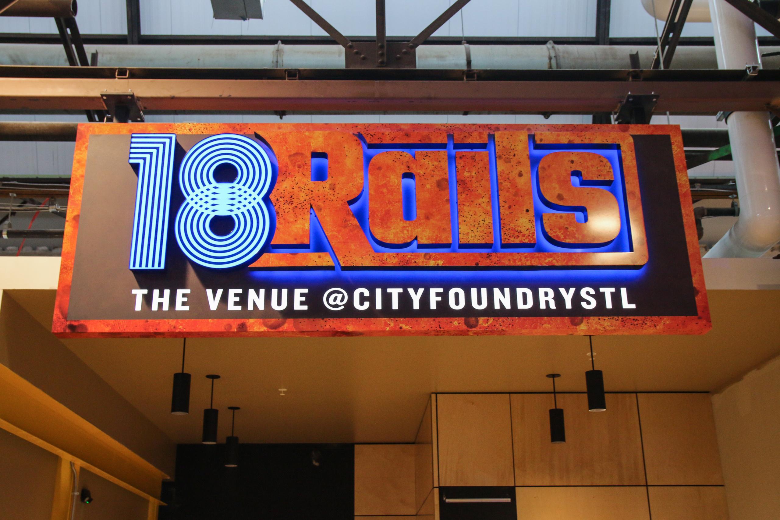 Sign reading "18 Rails: The Venue @ City Foundry STL" with colorful, stylized text, hanging from a ceiling in an industrial-looking setting.
