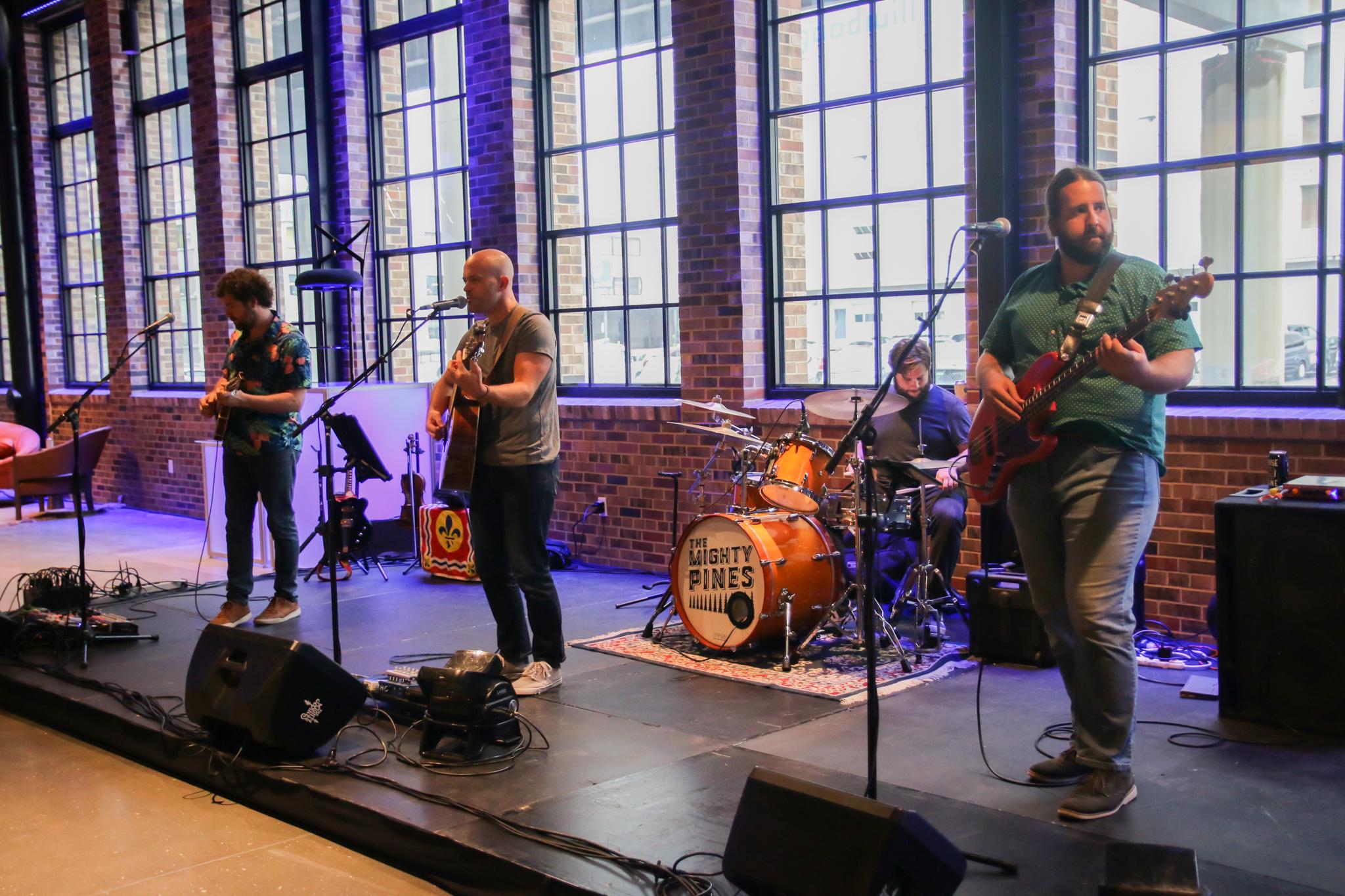 A band performs on stage with four musicians. One plays the drums, the others play guitars. They stand in front of large windows in a venue with brick walls. The drum set has "The Mighty Pines" written on it.