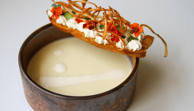 A rustic bowl filled with creamy white soup. Garnished above is a crispy crostini topped with whipped cream, green herbs, thin fried strips, and vibrant orange flowers.