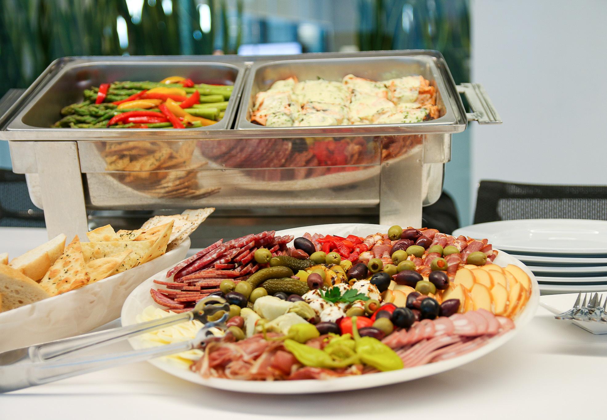 A buffet table featuring a platter of assorted deli meats, cheese, olives, and pickles, alongside a tray of roasted vegetables and what appears to be baked pasta. A basket of breadsticks and stack of plates are nearby.