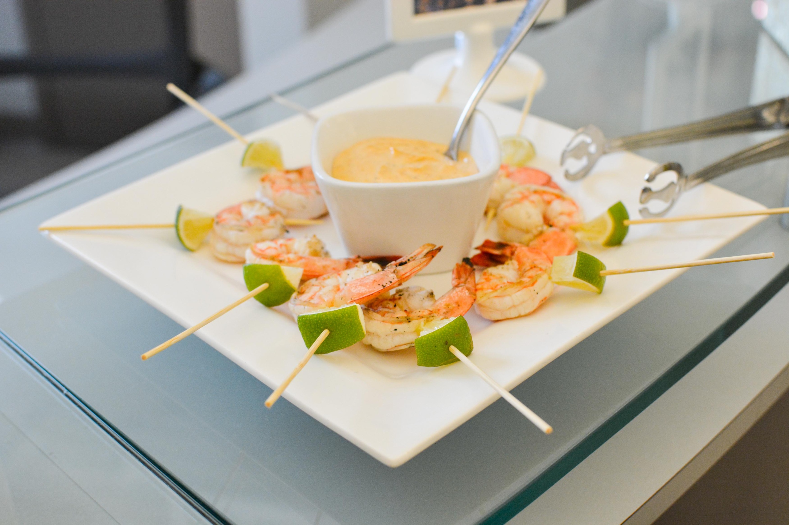 A square white plate with skewered shrimp and lime wedges arranged around a bowl of dipping sauce. Tongue and a utensil are placed beside the plate, set on a glass table.