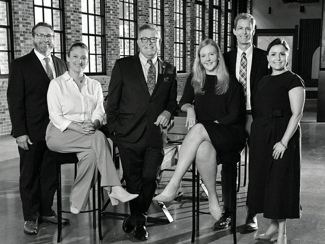 A group of six people, four men and two women, are posed together in a well-lit room with large windows. Two are seated on stools, and the rest are standing. They are dressed in professional attire, smiling at the camera. The image is in black and white.