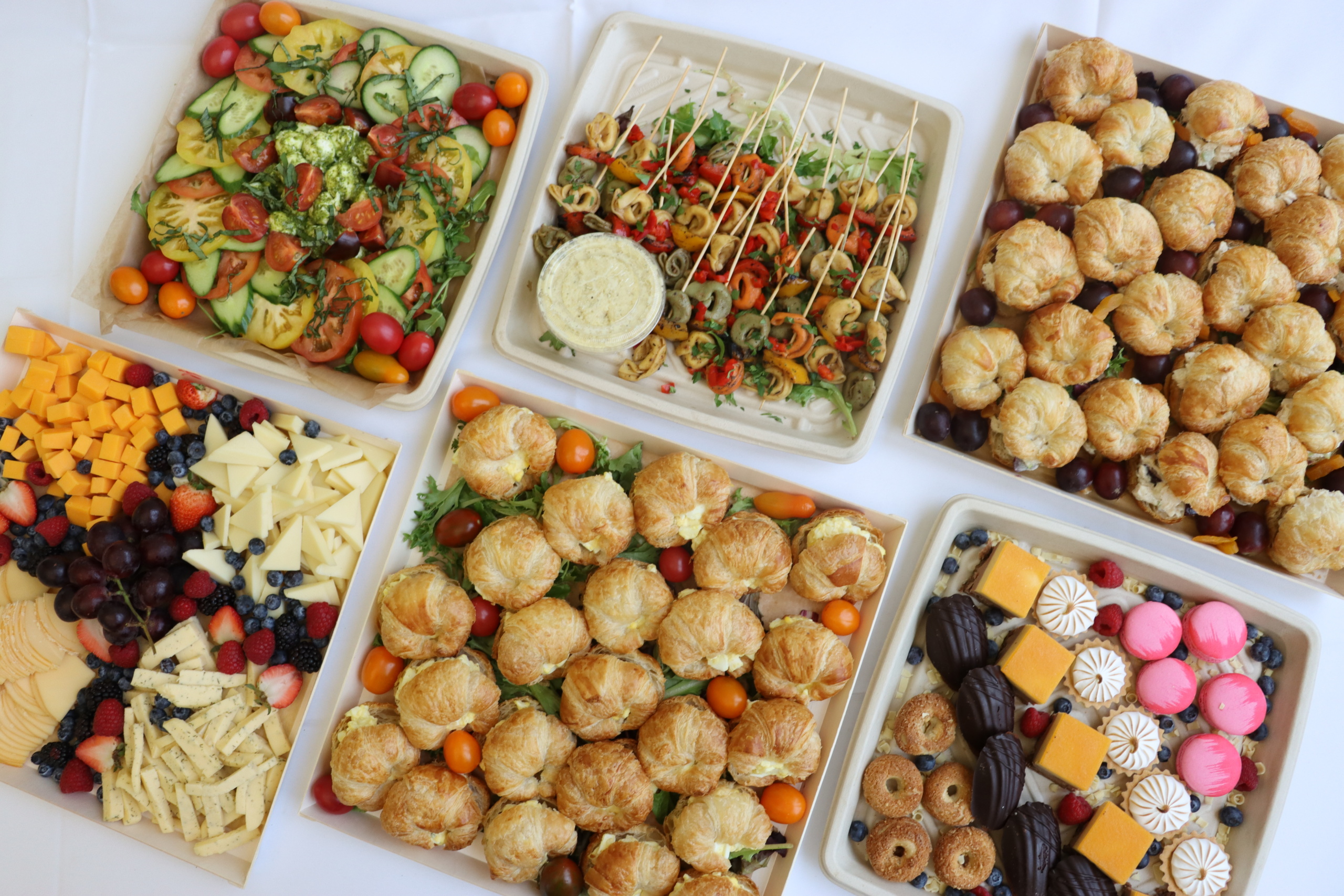 A spread of assorted finger foods arranged in trays. Includes a variety of sandwiches, cheese platter with fruits, mixed vegetables, pastry puffs, skewered appetizers with vegetables, and a tray of desserts with cookies and pastries.