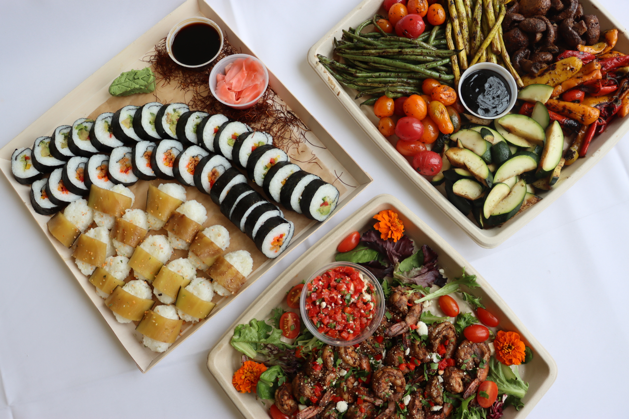Three platters of food on a table. One with sushi rolls and dipping sauce, another with grilled vegetables like asparagus and bell peppers, and a third with a salad topped with diced tomatoes and grilled shrimp. Decorated with flowers.