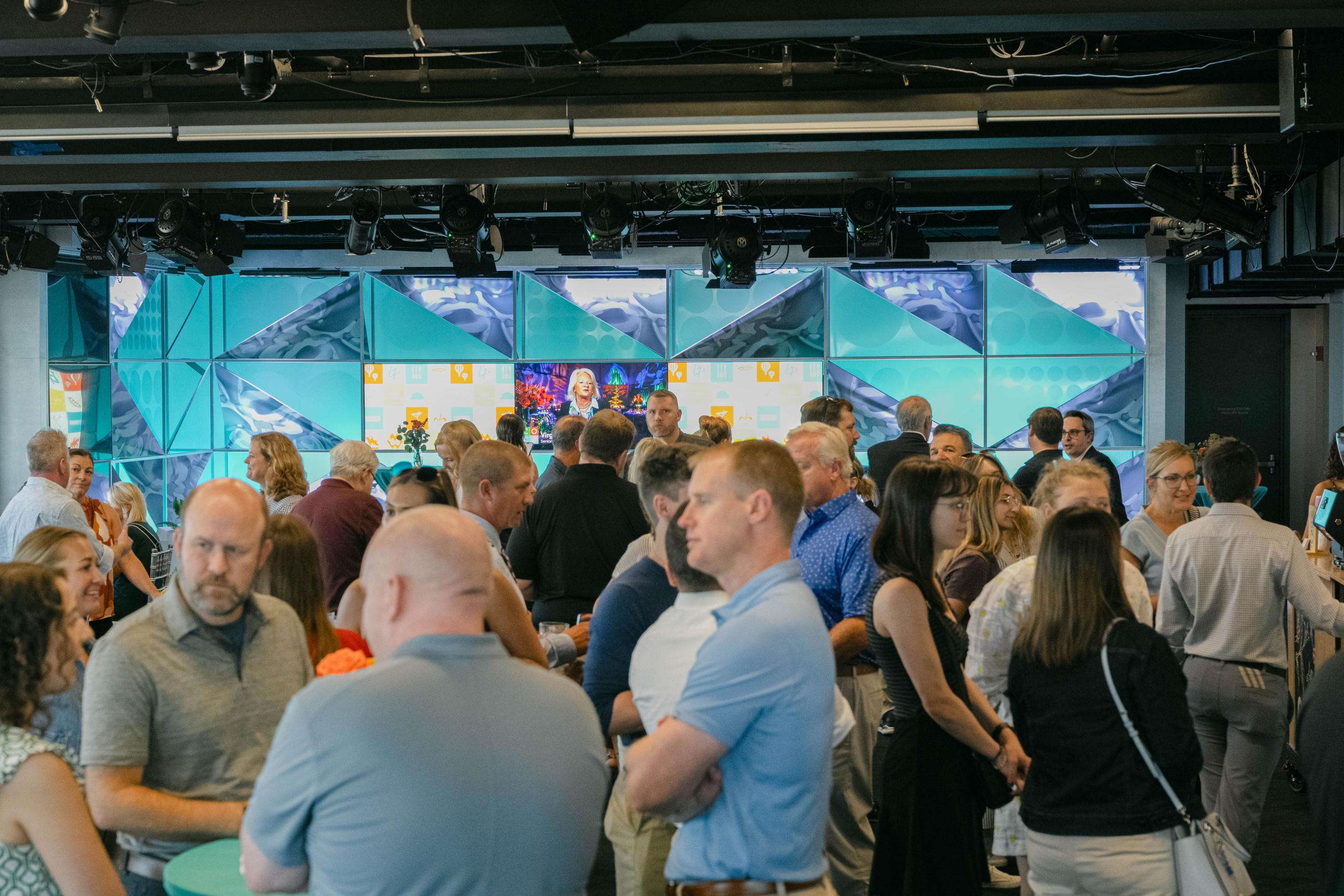 A crowd of people socializing at an indoor event with a large digital screen in the background.