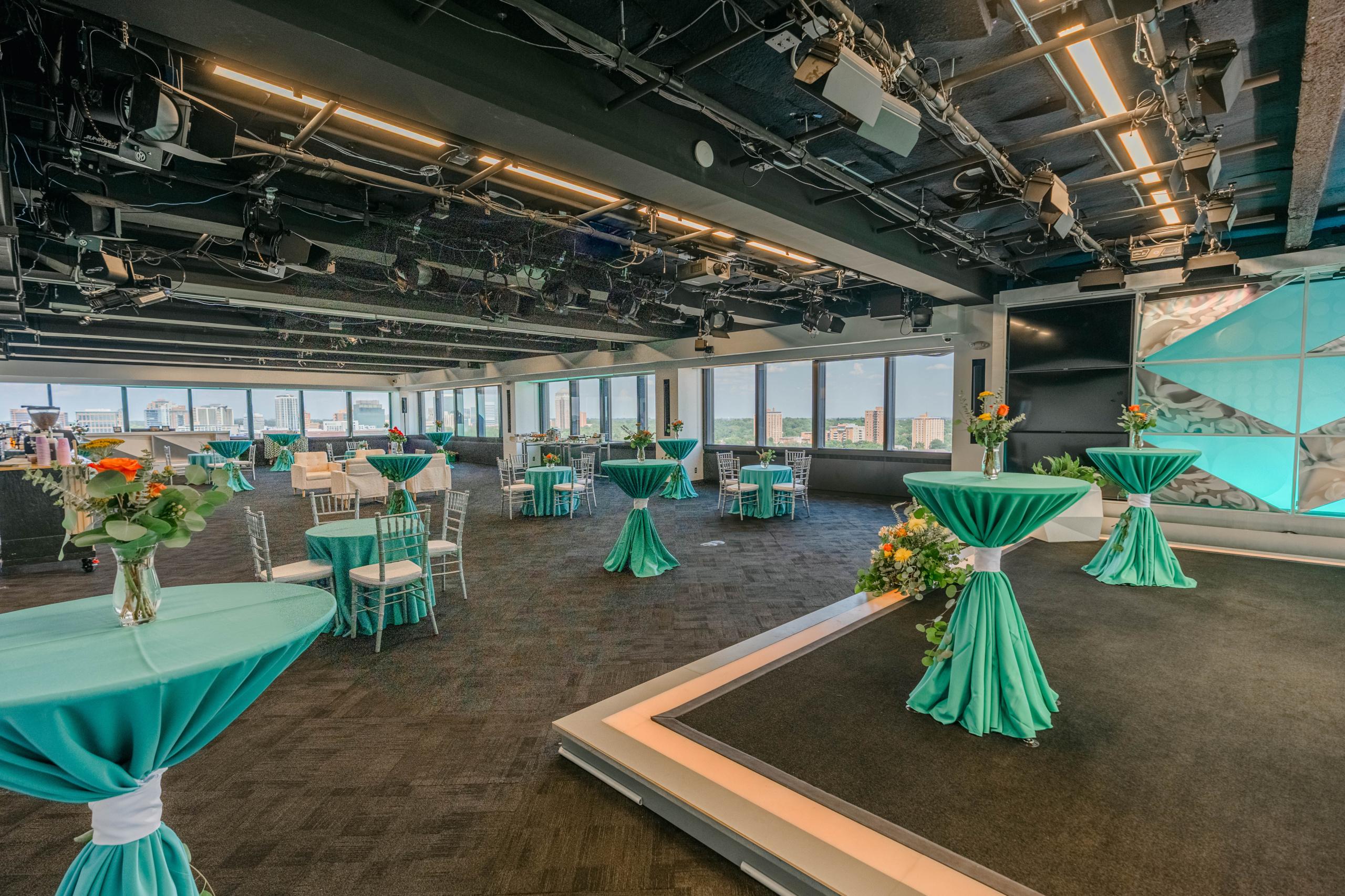 Event space featuring high-top tables with teal cloths, chairs, and a small stage.