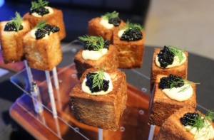 Close-up of gourmet appetizers on skewers: crispy toasted cubes topped with a dollop of creamy sauce, black caviar, and a sprig of dill. They are elegantly arranged on a clear display stand.