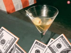 Martini glass with an olive, surrounded by stacks of 100-dollar bills on a table, with a striped background.