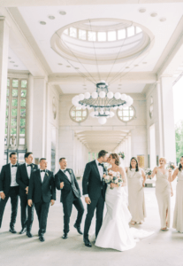 Wedding party celebrating as couple kisses under a grand chandelier in an elegant, open hallway.