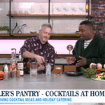Two men prepare cocktails in a modern kitchen studio with drinks and holiday snacks on the counter.